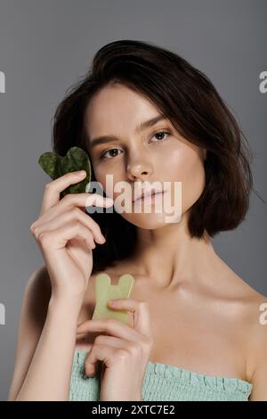 Une femme tient deux outils de jade gua Sha devant un fond gris. Banque D'Images