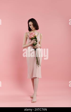 Une femme dans une robe rose se tient contre un fond rose vif, tenant un bouquet de fleurs roses. Banque D'Images