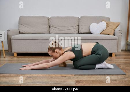 Une jeune femme pratique le yoga à la maison, agenouillée sur un tapis gris devant un canapé. Banque D'Images