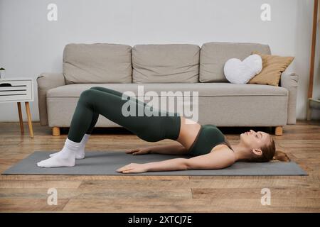 Une jeune femme en vêtements d'entraînement verts effectue une pose de pont sur un tapis de yoga dans son salon. Banque D'Images