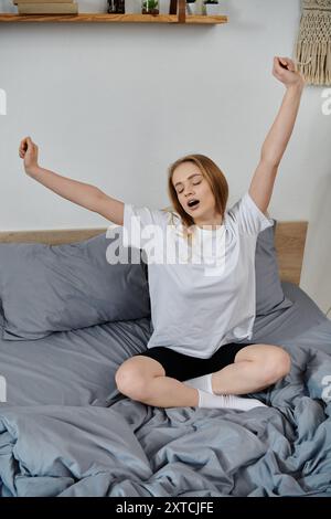 Une jeune femme s'assoit dans son lit et s'étire, bâillant alors qu'elle profite de la lumière du soleil du matin. Banque D'Images