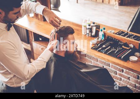 La coupe de cheveux doit être parfaite. Vue de dessus d'un jeune homme barbu se faisant couper les cheveux par un coiffeur tout en étant assis dans une chaise au salon de coiffure Banque D'Images