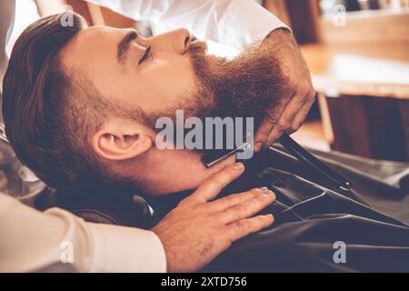 Tranchant et dangereux. Vue latérale rapprochée d'un jeune homme barbu se rasant avec un rasoir à bord droit par un coiffeur Banque D'Images