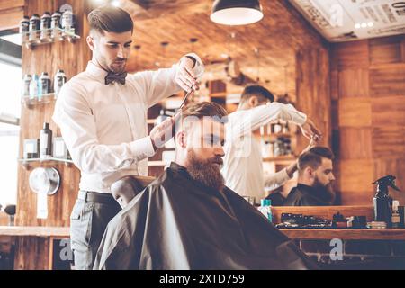 Tout devrait être parfait. Vue latérale d'un jeune homme barbu se faisant couper les cheveux par un coiffeur alors qu'il était assis dans une chaise au salon de coiffure Banque D'Images