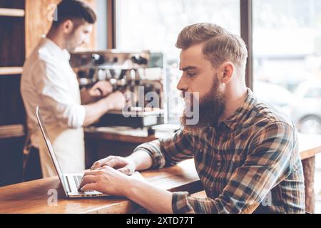 Travailler dans un café. Vue latérale d'un jeune homme barbu beau utilisant son ordinateur portable tout en étant assis au comptoir du bar au café avec le barista à l'arrière-plan Banque D'Images