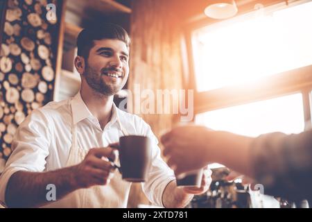 Voici votre café ! Gros plan sur une partie jeune homme beau dans un tablier donnant deux tasses à café à son client tout en se tenant debout au café Banque D'Images