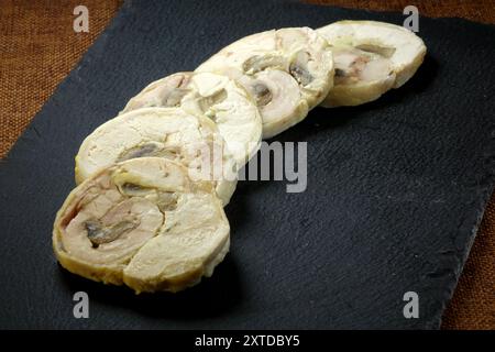 Un rouleau de viande magnifiquement arrangé coupé en tranches, garni de feuilles d'épinards frais et de graines de tournesol sur un tableau noir élégant. Banque D'Images