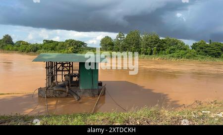 Une station de pompage flottante pour l'approvisionnement en eau brute, dérivant dans une rivière boueuse et teintée de rouge, avec une forêt dense et des nuages de tempête sombres qui se profilent dans le backgrou Banque D'Images