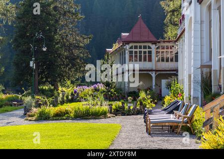 Une aile latérale de la Schatzalp à Davos présente un toit conique rouge au-dessus d'une véranda en bois blanchie et patinée. Un jardin de fleurs avec des vivaces violettes et jaunes s'étend devant. Des chaises longues rayées se dressent sur le chemin de gravier. Les architectes Otto Pfleghard et Max Haefeli ont construit le sanatorium contre la tuberculose entre 1898 et 1900 pour le pionnier néerlandais de Davos Willem Jan Holsboer, qui l'a conçu comme un centre de santé progressiste. L'ancien sanatorium est aujourd'hui un hôtel. Thomas-Mann-Weg, Davos, Grisons, Suisse. Banque D'Images