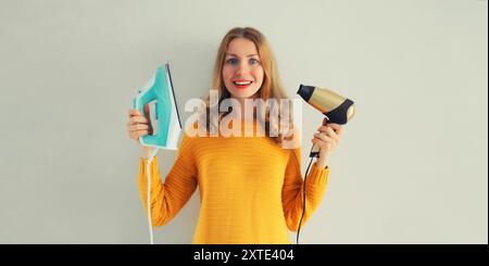 Tâches ménagères, repassage - femme au foyer heureuse souriante tenant le fer et le sèche-cheveux dans ses mains sur le mur gris à la maison Banque D'Images