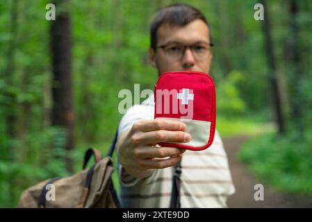 Randonneur dans la forêt tient une trousse de premiers soins rouge vers la caméra, mettant l'accent sur la préparation et la sécurité pendant les aventures en plein air. Banque D'Images