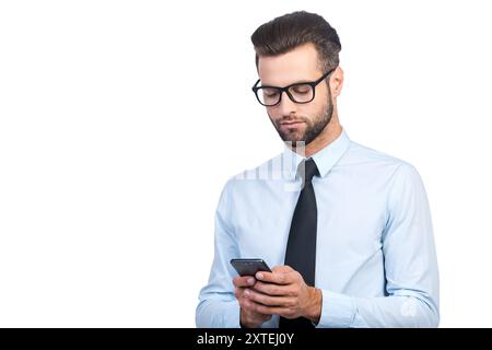 Saisie d'un message professionnel. Confiant jeune homme beau en chemise et cravate tenant le téléphone portable et le regardant tout en se tenant debout sur fond blanc Banque D'Images