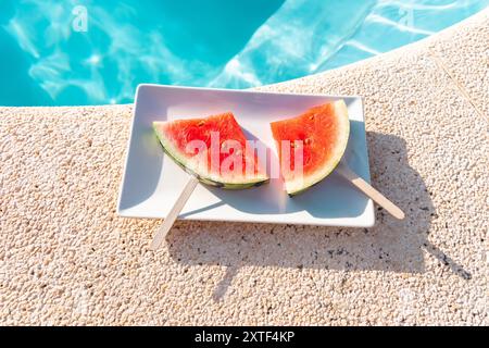 Tranches de pastèque rafraîchissantes sur des bâtonnets placés sur une assiette au bord d'une piscine bleu vif Banque D'Images