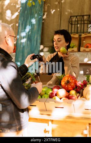 Propriétaire d'une petite entreprise agricole se tient derrière le comptoir, vendant des fruits et légumes biologiques frais. Une vendeuse et une cliente senior parlent de produits biologiques sains cultivés localement. Banque D'Images