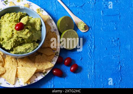 Version brésilienne de guacamole dans un bol bleu avec une tomate cerise sur le dessus entouré de nachos sur le dessus d'une table bleue. Banque D'Images