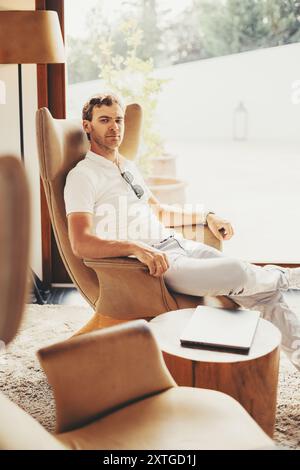 Un homme portant une chemise blanche et un pantalon s'assoit décontracté dans un fauteuil moderne beige, avec son bras gauche reposant sur l'accoudoir de la chaise et un ordinateur portable placé sur un Banque D'Images