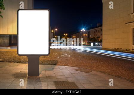 Maquette vierge du panneau d'affichage vertical d'affiche de rue sur fond de ville la nuit. Visionneuse publicitaire devant les allées des feux de voiture Banque D'Images