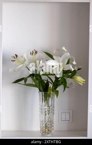 Un vase en verre transparent rempli de feuilles vertes et de lis ouverts, placé sur une étagère blanche contre un mur blanc Uni. Banque D'Images