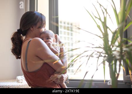 Femme regarde par la fenêtre de sa maison tout en tenant sa fille nouveau-née dans ses bras. Banque D'Images