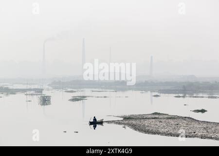 Un pêcheur dans un bateau de campagne attend patiemment sa prise le long de la rivière hazy Turag à Asulia au Bangladesh. La zone est remplie de fours de briques à cheminée fixe, qui viennent de transformer leur production en grande vitesse. Pendant la plus grande partie de l'année, les fours en briques sont inondés car ils sont situés sur les lits de la rivière. Lorsque l'eau recule pendant la saison sèche, les fours reviennent à la vie alors que des centaines d'ouvriers empilent des briques fraîchement pressées en silt dans d'énormes fours à bois et à charbon. En utilisant une technique simple qui remonte à des centaines d'années, les fours fixes de brique de cheminée fournissent un approvisionnement constant Banque D'Images