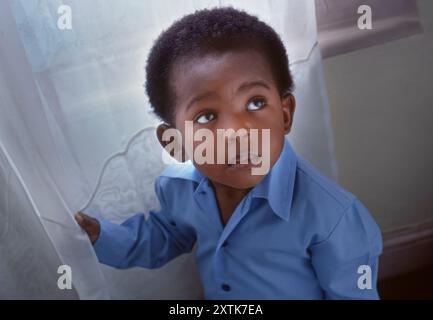 Bébé de 4 ans Noir afro-américain garçon près de la fenêtre à la maison seul regarde solennel et pensif, mais très mignon beau et attrayant, éclairé seulement par la lumière naturelle de la fenêtre. Banque D'Images