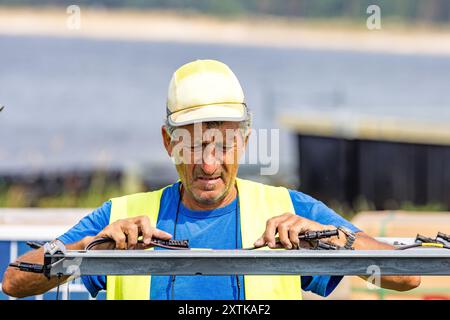 Neuendorf, Allemagne. 15 août 2024. Un ouvrier de Pfalzsolar monte des modules solaires pour le système photovoltaïque flottant sur la mer Baltique de Cottbus. Une fois terminé, il couvrira 18 hectares du lac (environ un pour cent) avec 51 000 modules solaires. En moyenne, le système peut générer 290000 mégawattheures d'énergie par an. Les corps flottants avec les modules solaires attachés à eux sont reliés au lit du lac par 34 points d'ancrage de sorte que les fluctuations des niveaux d'eau peuvent être compensées. Crédit : Frank Hammerschmidt/dpa/Alamy Live News Banque D'Images