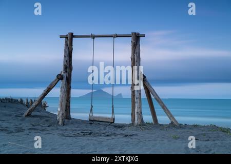 Une balançoire solitaire en bois vous attend sur une plage tranquille. Au loin, l'île de la tortue est partiellement voilée par des nuages, créant un l côtier serein Banque D'Images