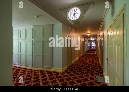 La Schatzalp au-dessus de Davos, Grisons, montre un couloir avec un tapis à motifs rouges et des placards encastrés crème dans le bâtiment principal. Une grande horloge double face avec un cadran blanc est suspendue dans le couloir. Otto Pfleghard et Max Haefeli ont construit l’ancien sanatorium entre 1898 et 1900 pour Willem Jan Holsboer. Des portes supplémentaires de chambres d’hôtes et un coin salon recouvert d’un tapis rouge sont visibles à l’extrémité du couloir, Bobbahnstrasse, Suisse. Banque D'Images