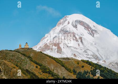 Gergeti Trinity Church perchée sur une colline avec le mont Kazbek enneigé dominant en arrière-plan, sous un ciel bleu vibrant en Géorgie. Banque D'Images