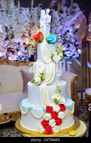 Un gâteau de mariage à quatre niveaux décoré de roses blanches et bleues, avec deux statues de jeunes mariés sur le dessus Banque D'Images