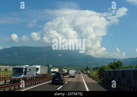 Ossenigo, Italie - 8 juin 2023 : les voitures voyagent le long d'une route panoramique entourée de montagnes majestueuses et d'un ciel bleu vif rempli de nuages blancs gonflés. Banque D'Images