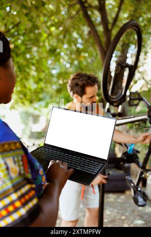 Jeune femme afro-américaine utilisant un ordinateur portable avec écran isolé faisant l'entretien du vélo à la cour de la maison. Mini-ordinateur affichant un écran vierge pendant que deux amateurs de sport actifs réparent une bicyclette cassée. Banque D'Images