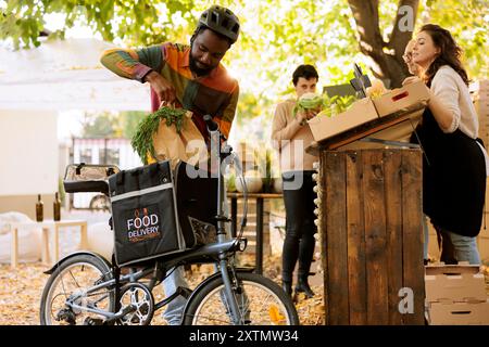 Black Man met des produits bio biologiques dans un sac thermique, prend une commande au marché des agriculteurs et fait du vélo. Courrier africain livrant des fruits et légumes frais de la ferme aux clients. Banque D'Images