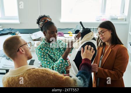 Jeune couturière afro-américaine coudre le col à la blouse blanche sur mannequin lors du travail d'équipe avec deux autres tailleurs Banque D'Images