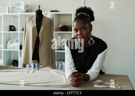 Jeune tailleur afro-américaine ou créatrice de mode regardant la caméra tout en se tenant debout près de la table avec du textile gris déroulé Banque D'Images