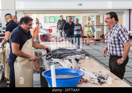 Deux hommes parlent au-dessus d'un comptoir alors que le poissonnier utilise son couteau pour préparer un poisson noir Scabbard au marché Mercado dos Lavradores à vendre. Illustré à la Banque D'Images