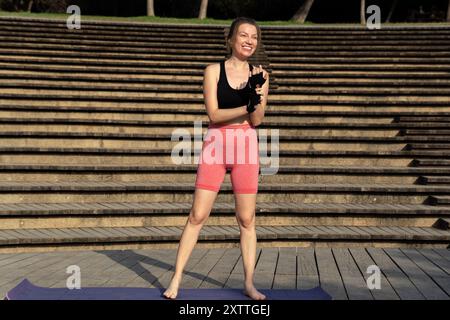 Une jeune femme aux cheveux roux sourit brillamment, debout sur une terrasse en bois au soleil, vêtue de vêtements de sport et de gants, prête pour son cours de yoga pilates Banque D'Images