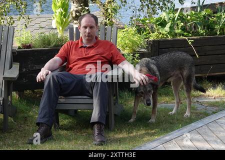 Un homme sérieux caresse son chien mongrel. Il est assis sur une chaise de jardin. Derrière lui un potager Banque D'Images