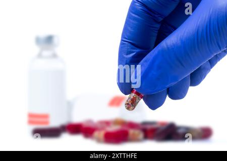 Main du médecin tenant la pilule rouge dans des gants bleus devant des flacons médicaux de vaccin et des pilules avec le focus out sur fond blanc Banque D'Images