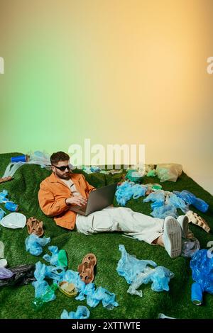 Un homme est allongé sur une pelouse artificielle entourée de déchets plastiques, travaillant sur un ordinateur portable. Banque D'Images