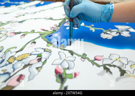 Un artisan habile portant un gant blanc peint délicatement un motif floral bleu foncé sur une écharpe de soie blanche, exposée sur une table en bois, mettant en valeur met Banque D'Images