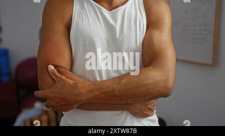 Jeune homme hispanique dans un débardeur blanc debout à l'intérieur dans une salle de gym avec les bras croisés montrant les bras musclés Banque D'Images