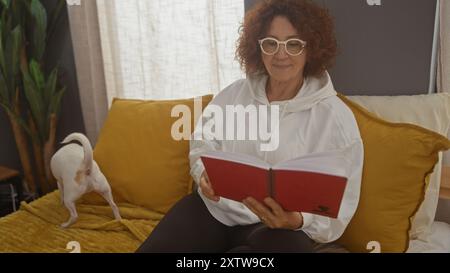 Une femme âgée lit un livre sur un lit confortable dans une chambre confortable tandis que son chien chihuahua se tient à proximité. Banque D'Images