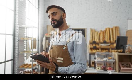 Jeune homme hispanique barbu utilisant une tablette à l'intérieur dans une boulangerie avec des étagères de pain frais et de pâtisseries en arrière-plan Banque D'Images