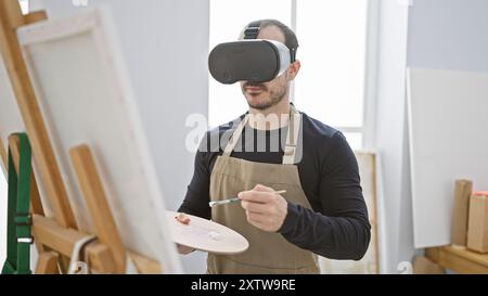 Homme dans un tablier utilisant un casque vr tout en peignant sur toile dans un intérieur de studio lumineux. Banque D'Images