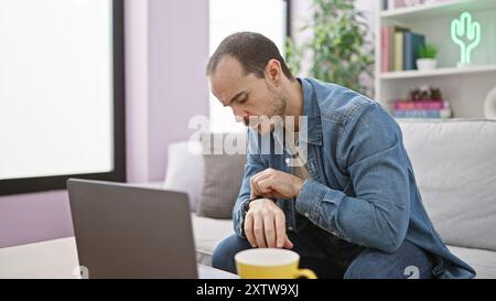 Homme hispanique avec la barbe vérifiant le temps sur sa montre-bracelet à l'intérieur, assis sur un canapé de salon. Banque D'Images