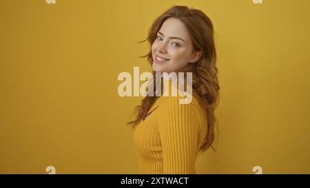 Une belle jeune femme aux cheveux blonds sourit tout en se tenant debout sur un mur de fond jaune isolé, mettant en valeur son style vibrant et son deme joyeux Banque D'Images