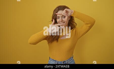 Une belle femme blonde portant un pull jaune pose de manière ludique sur un fond jaune isolé, encadrant son visage avec ses mains. Banque D'Images