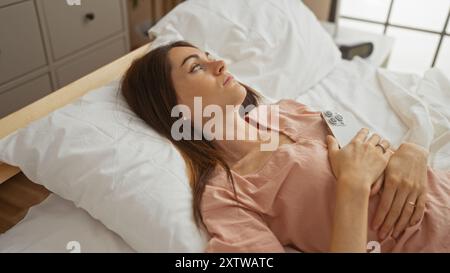Femme allongée sur le lit dans une chambre tenant un smartphone à l'air réfléchi à l'intérieur avec la lumière naturelle et des oreillers blancs montrant l'atmosphère paisible de la maison Banque D'Images