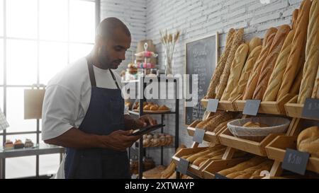 Jeune homme travaillant dans une boulangerie, portant un tablier et utilisant une tablette entourée de différents types de pain. Banque D'Images